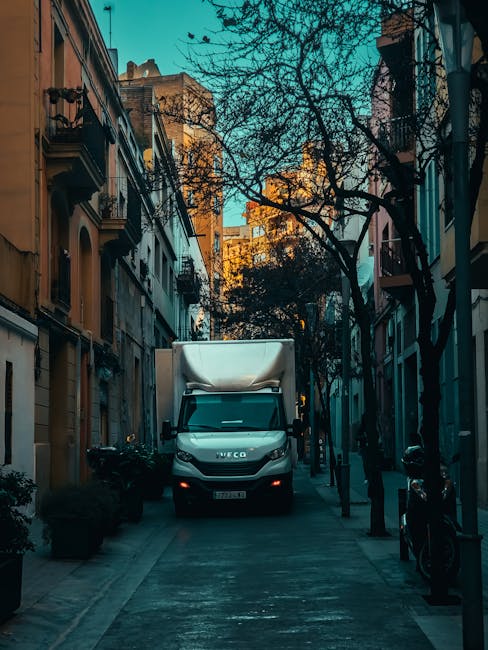 A white Iveco moving van is parked on a narrow residential street with multi-storey buildings on either side. The van's rear doors are closed, and it is positioned centrally in the image, indicating an active loading or unloading process. The street is lined with leafless trees and masts of bollards along the pavement, suggesting a typical urban environment. The surrounding buildings feature balconies, windows, and brick or stucco facades, with some shadows cast by the late afternoon sunlight. The scene reflects a home relocation setting, with the van ready for furniture transport as part of a professional removals service by Man and Van Belmont. Overall, the image captures a straightforward moment during packing and moving activities, highlighting the urban logistics involved in house removals.