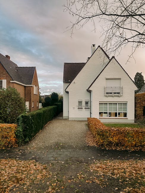 A white two-storey house with a pitched roof and a small front garden area, located on a calm residential street. The house features a large ground-floor window with multiple panes, and a small balcony with a metal railing on the first floor. To the left of the house, there is a paved pathway leading from the street to the front door, bordered by neatly trimmed hedges with orange and green foliage. The surrounding area includes other houses with brick and tiled exteriors, some trees with sparse branches, and a cloudy sky overhead. This scene depicts a typical suburban setting suitable for home relocation or furniture transport, with the house prepared for a moving process by a professional removals service such as Man and Van Belmont, which specialises in residential removals and packing services.
