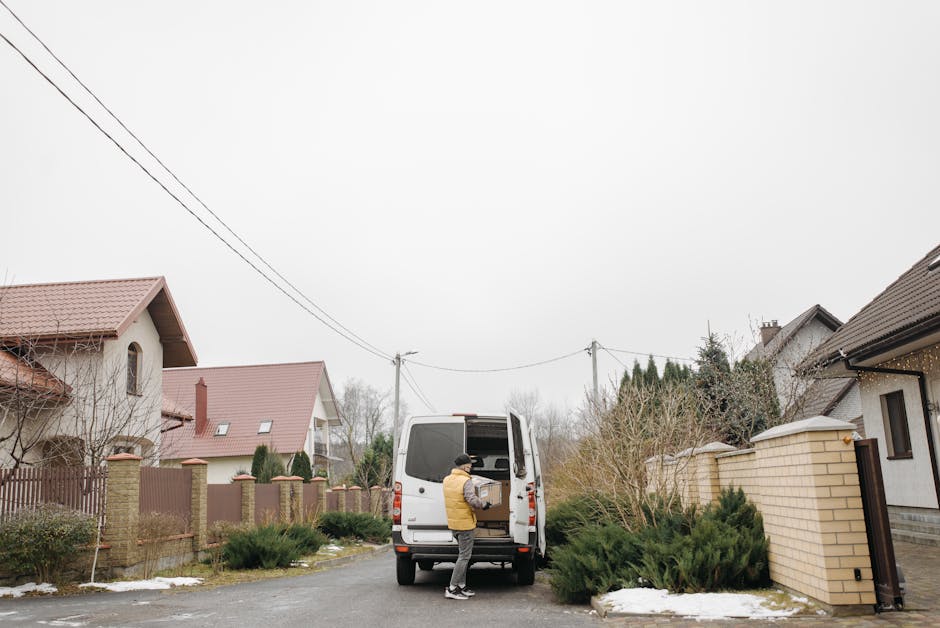 A person dressed in a yellow jacket and black hat loading a cardboard box into the back of a white van on a residential street, surrounded by houses with pitched roofs, brick and wooden fences, and bushes. The van is parked on the driveway of a house, with other belongings possibly inside. Overhead power lines run across the cloudy sky. This scene depicts the process of home relocation, furniture transport, or packing and moving activities, with the vehicle positioned for efficient loading on a narrow, tight driveway often encountered during house removals by Man and Van Belmont. The environment shows winter conditions with some snow on the ground and shrubbery, and the overall setting emphasizes moving logistics within a suburban neighbourhood.