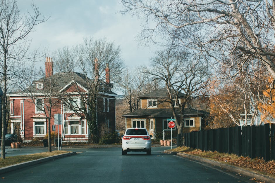 A white two-storey house with a pitched roof and a small front garden area, located on a calm residential street. The house features a large ground-floor window with multiple panes, and a small balcony with a metal railing on the first floor. To the left of the house, there is a paved pathway leading from the street to the front door, bordered by neatly trimmed hedges with orange and green foliage. The surrounding area includes other houses with brick and tiled exteriors, some trees with sparse branches, and a cloudy sky overhead. This scene depicts a typical suburban setting suitable for home relocation or furniture transport, with the house prepared for a moving process by a professional removals service such as Man and Van Belmont, which specialises in residential removals and packing services.