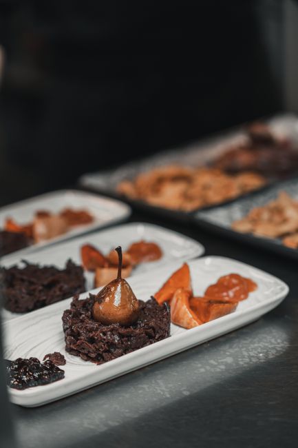 The image shows a white rectangular serving platter with three portions of rich chocolate brownies, each topped with a whole cherry and a small chocolate stick. To the right of the brownies, there are several slices of toasted orange-colored dried fruit, possibly apricots or peaches. In the background, slightly out of focus, are additional similar plates with more baked desserts and dried fruit slices, placed on a dark grey or black countertop surface. The scene appears to be set in a professional kitchen or dessert presentation area, with ambient lighting highlighting the textures and colors of the food items. The overall composition emphasizes the arrangement and variety of chocolate and dried fruit treats, suitable for a catering or event setting.