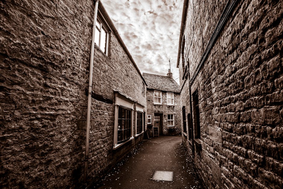 A narrow alleyway between two stone-built houses with textured brick and rough sandstone walls. The alley is paved with asphalt and appears slightly curved, with small white scattered debris on the ground. On the left side, there are three white-framed windows with grid-patterned panes, part of a building that extends partly into the scene. On the right, another stone wall features a small window and a black drainpipe running vertically. At the end of the alley, a small courtyard with a building that has multiple windows, including a set of double-hung sash windows, is visible under an overcast sky with cloud cover. The scene depicts a typical British residential street, suitable for house removals and furniture transport services, with visible details of the building materials and environment where loading and packing activities might take place, as handled by companies like Man and Van Belmont.