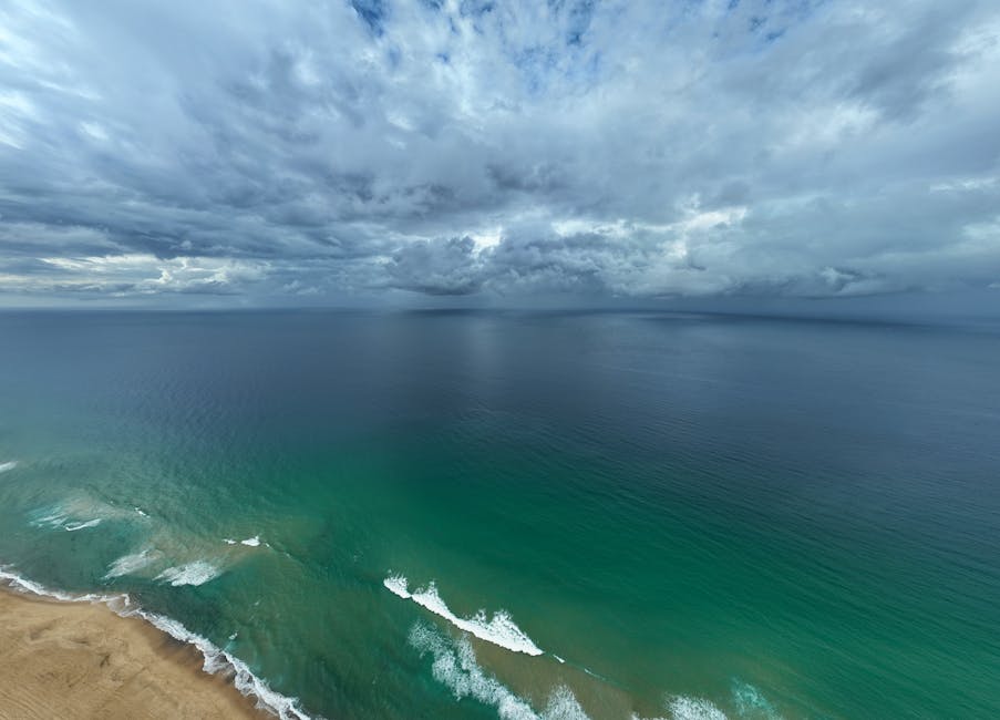 Aerial view of a coastal scene showing a sandy beach in the foreground with gentle waves breaking along the shoreline. Beyond the beach, the calm, deep blue sea extends to the horizon under a vast, cloudy sky filled with dark and light grey clouds. The overcast weather creates a slightly moody atmosphere, with subtle light filtering through the clouds. The image captures the natural landscape before any house removals or moving activities, focusing on the open sea and coastal environment. Such scenery is often encountered during travel or transport to a seaside home location, which may relate to the services provided by Man and Van Belmont for home relocation, furniture transport, and moving logistics. The tranquil yet expansive view emphasizes the scale of the area involved in house removals and the importance of careful planning during the loading process and transport of household items.
