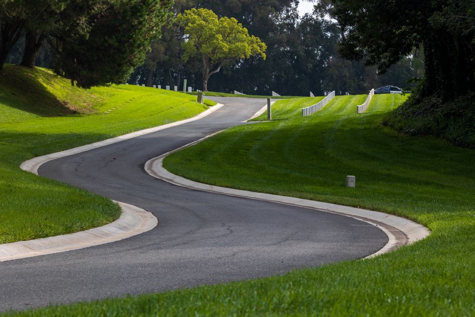 A winding residential driveway with smooth dark asphalt surface, bordered by neatly maintained green grass and curved concrete edges. The driveway features gentle curves and slight elevation changes, with white wooden fences lining the sides and tall leafy trees providing shade in the background. A black car is parked at the top of the drive near a small white post. The scene is well-lit with natural sunlight, highlighting the lush greenery and the clean, organized environment. This setting illustrates the typical layout encountered during house removals or home relocation projects, where careful navigation of narrow or curved drives is necessary for furniture transport and loading processes, as handled by companies like Man and Van Belmont.