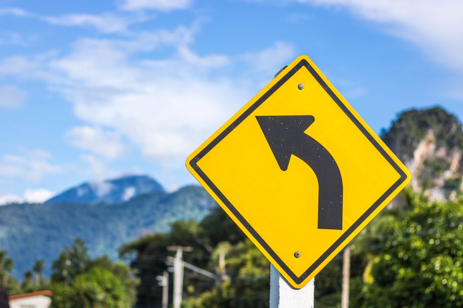 A yellow diamond-shaped road sign with a black leftward curve arrow stands prominently outdoors against a backdrop of a mountain landscape with green trees, a partly cloudy sky, and distant rocky peaks. The sign is mounted on a metal pole situated on a paved area near a property entrance, possibly indicating a sharp bend or narrow turn on Belmont Road during a home relocation or furniture transport process. The environment includes visible utility poles and overhead wiring, suggesting a residential or semi-rural setting. This image, representing common challenges faced during house removals along tight or winding roads, is relevant to the services provided by Man and Van Belmont, especially when planning moving logistics and navigating narrow turns during house removals or packing and loading activities.