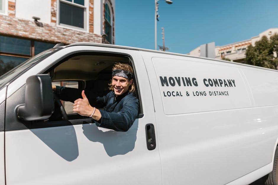 A smiling young man wearing a black bandana and a dark blue shirt is sitting in the driver’s seat of a white moving van parked outside a building. He is giving a thumbs-up gesture while looking out of the open window. The side of the van displays the text 'MOVING COMPANY' followed by 'LOCAL & LONG DISTANCE' in bold black letters, indicating a professional house relocation service. The vehicle is positioned on a paved street with a clear blue sky overhead, and nearby structures include a brick building with large windows and another modern structure in the background. The scene captures the loading process or transit phase of furniture transport and packing during a home move, with equipment such as the van and the driver's readiness suggesting the start or ongoing part of a house removal. Man and Van Belmont occasionally offer such services as part of their removals category, ensuring professional moving logistics.