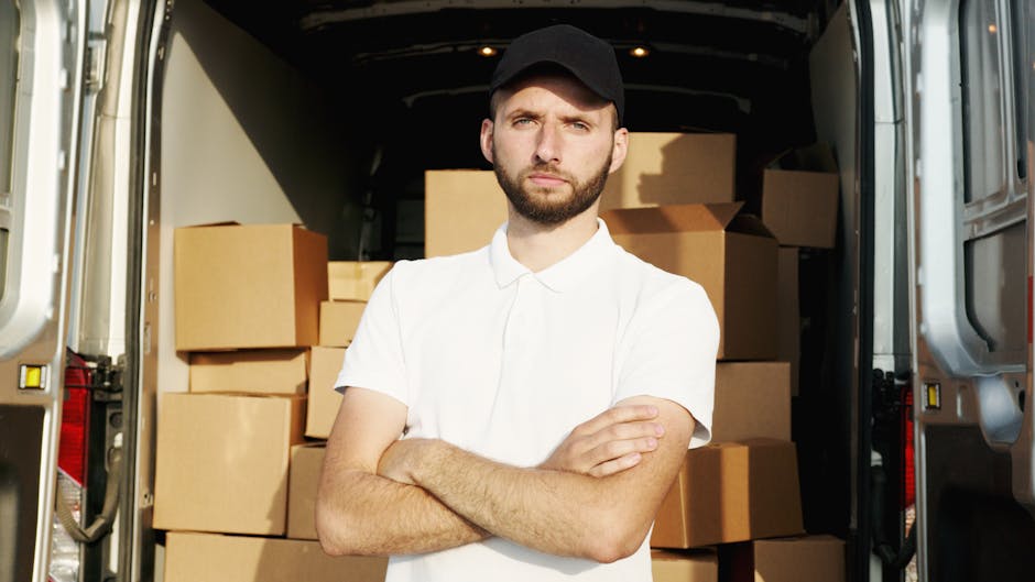 A man standing inside the open rear of a moving van during a home relocation process, with multiple packed cardboard boxes of various sizes stacked behind him. The boxes are sealed with packing tape, some with handles visible, and are arranged in an organized manner to facilitate loading. The man, wearing a white polo shirt and a black cap, has a serious expression with arms crossed, indicating a professional approach to furniture transport and packing and moving logistics. The interior of the van is dark with some ambient lighting, and part of the vehicle's metallic exterior is visible on the sides. The scene captures the loading process, highlighting the importance of careful packing and proper organisation in successful removals, as experienced by companies like Man and Van Belmont in Belmont during house removals.