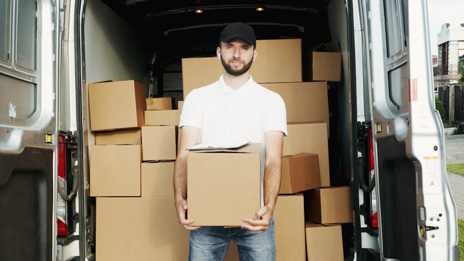 A smiling young man wearing a black bandana and a dark blue shirt is sitting in the driver’s seat of a white moving van parked outside a building. He is giving a thumbs-up gesture while looking out of the open window. The side of the van displays the text 'MOVING COMPANY' followed by 'LOCAL & LONG DISTANCE' in bold black letters, indicating a professional house relocation service. The vehicle is positioned on a paved street with a clear blue sky overhead, and nearby structures include a brick building with large windows and another modern structure in the background. The scene captures the loading process or transit phase of furniture transport and packing during a home move, with equipment such as the van and the driver's readiness suggesting the start or ongoing part of a house removal. Man and Van Belmont occasionally offer such services as part of their removals category, ensuring professional moving logistics.