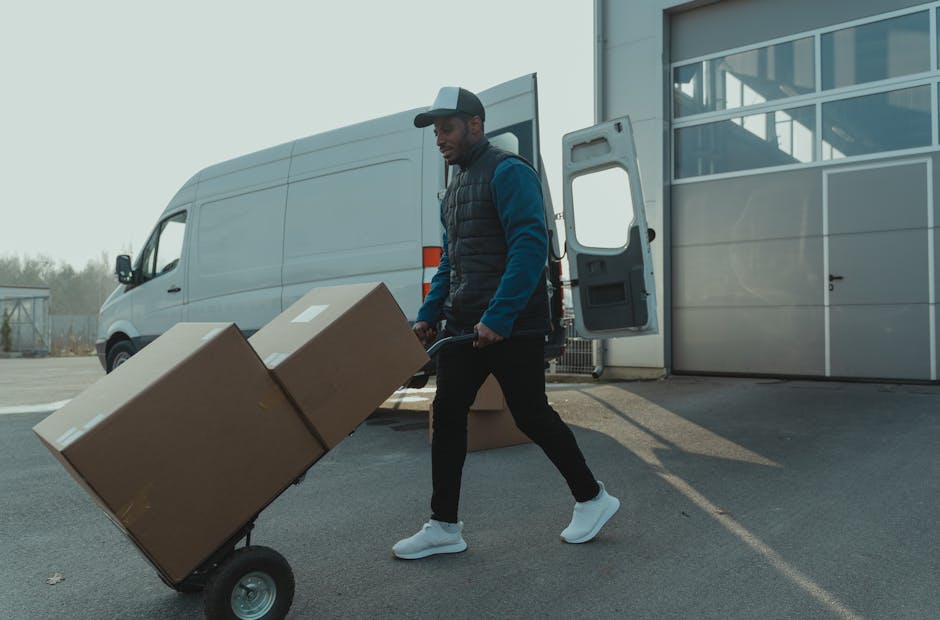 A man wearing a black cap, blue jacket, and black trousers is walking outside a modern building with glass windows, pushing a hand truck loaded with two large cardboard boxes wrapped in clear plastic. The boxes are secured with tape and stacked securely on the trolley. Behind him, a white moving van with its sliding door open is positioned on a concrete driveway, indicating an ongoing relocation process. The van's interior is partially visible, showing some packing materials and additional boxes inside. The scene is illuminated by natural daylight, and there are some leafless trees and a fence in the background, suggesting a residential or commercial property setting. This image reflects furniture transport and packing activities as part of home relocation or moving services provided by Man and Van Belmont, which specializes in last-minute moves and removals.