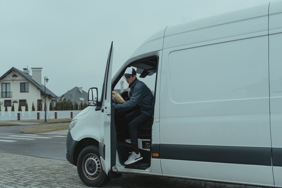 A man wearing a black baseball cap, glasses, a dark jacket, and black pants is seated in the driver's seat of a white cargo van, with the door open, on a residential street. He is holding a cardboard box, preparing to load it into the vehicle as part of a house removal process. Inside the van, there are visible packing materials and boxes, with additional packed items on the pavement nearby. The van is parked on the street in front of a house with a tiled roof, white exterior walls, and a garden surrounded by a white picket fence. In the background, there are other similar houses, a lamppost, and a crosswalk across the road. The scene is under natural daylight, with overcast skies, capturing a typical home relocation or furniture transport operation as seen during packing and loading steps, performed by Man and Van Belmont, specialising in removals.