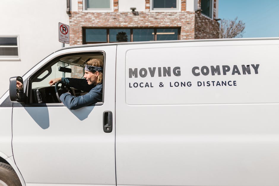 A male removal driver with light brown hair, wearing a black bandana and a dark jacket, is seated inside a white moving van, visible through the open front passenger door. The van is parked on the street in front of a brick residential building with large windows. The side of the van displays the text 'MOVING COMPANY' in large bold letters, with the phrases 'LOCAL & LONG DISTANCE' underneath. Inside the van, there are packed cardboard boxes and furniture items covered with protective blankets, indicative of a house relocation process. The area around the van is clear, and the environment appears bright with natural daylight, suggesting daytime. The scene captures the loading process typical of professional removals services, such as those offered by Man and Van Belmont, as part of their house removals and furniture transport operations, aligned with Sutton Council permit rules for Belmont moving vans.