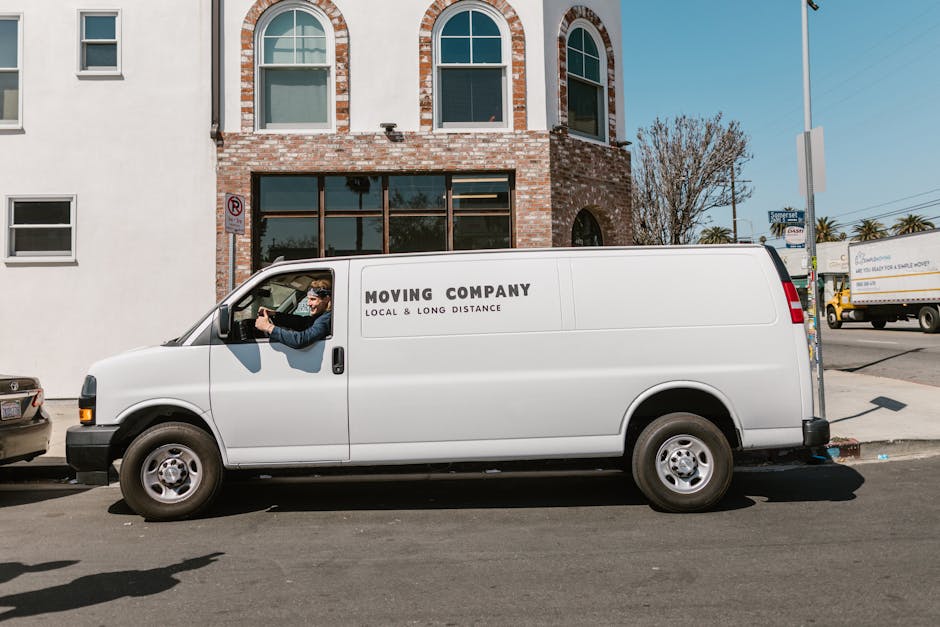 A white moving van with 'MOVING COMPANY LOCAL & LONG DISTANCE' printed on the side parked on a street in front of a brick and white building with arched windows. The driver, wearing sunglasses and a dark jacket, is seated inside, with one arm resting on the open window and looking towards the camera. The van is positioned near the pavement with a small pile of packing materials, such as cardboard boxes and blankets, visible inside. Behind the van, other vehicles, including a small car and a larger truck, are visible on the road. The scene is well-lit with clear weather, and nearby trees and street signs indicate an urban environment. This image depicts furniture transport and home relocation activities carried out by a professional removals service, with typical moving equipment seen around the vehicle, supporting the logistics of packing and loading household items.
