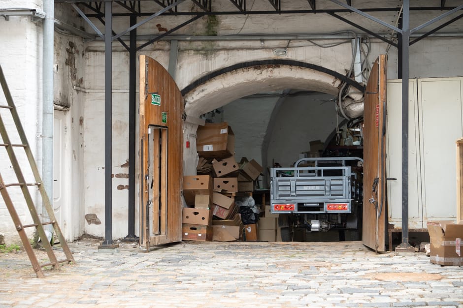 Photograph showing a rear loading area of a property during a home relocation process, with open wooden double doors revealing a small flatbed truck parked inside. The truck is partially loaded with cardboard boxes and packing materials, including some boxes that appear to be stacked and others scattered around. To the left, a wooden ladder leans against the wall leading up to the building, which has a cobbled stone ground outside. The interior behind the open doors includes a white-washed arched ceiling and visible pipes, indicating a storage or utility space. The scene is lit with natural light, highlighting the boxes, truck, and surrounding environment. As part of the furniture transport and packing process, Man and Van Belmont is assisting with the logistics of moving household items through this access point, ensuring proper handling during the loading process for a local house removal.